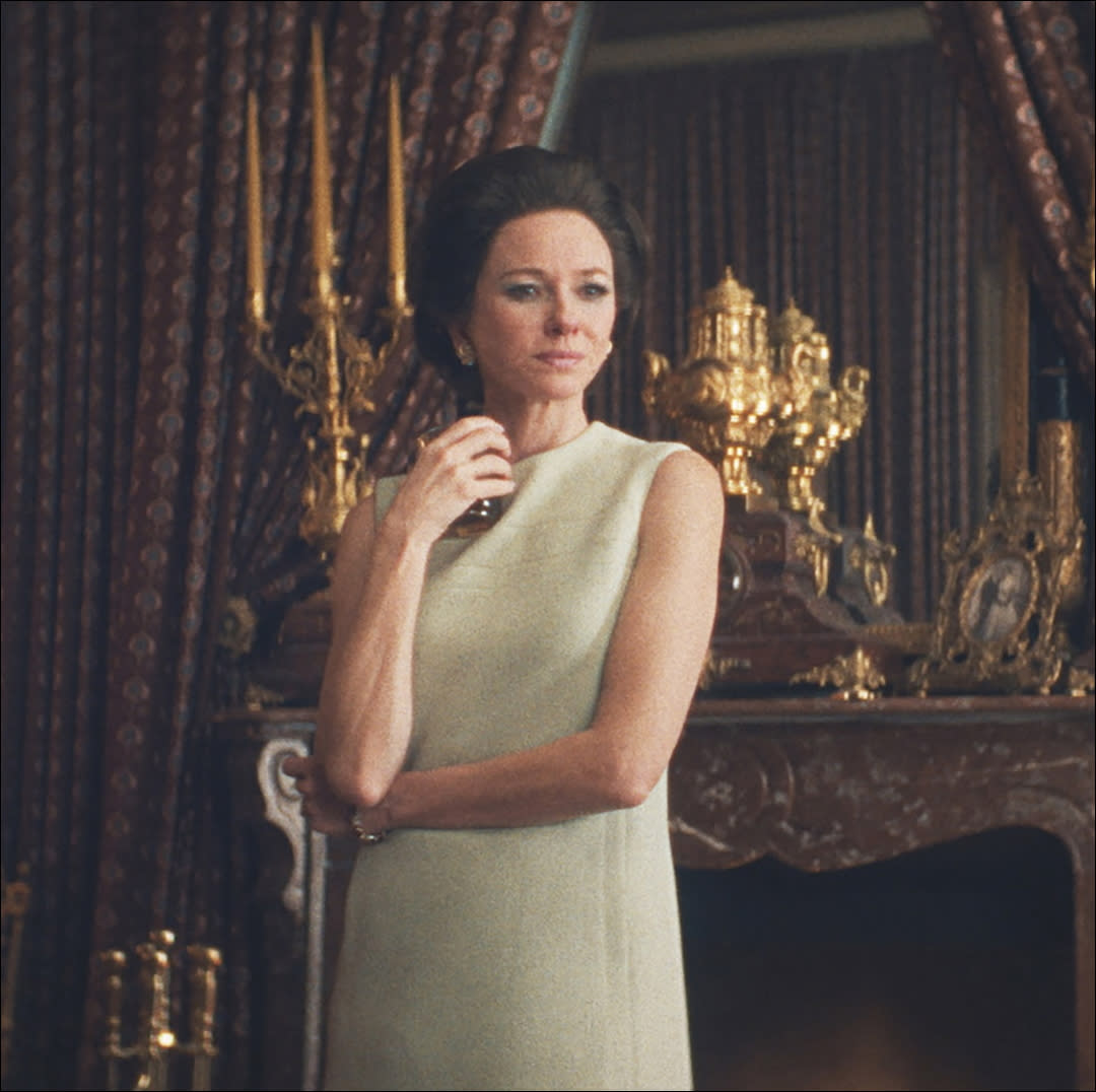  A woman (naomi watts as babe paley) stands in front of an ornate fireplace and candelabras. 