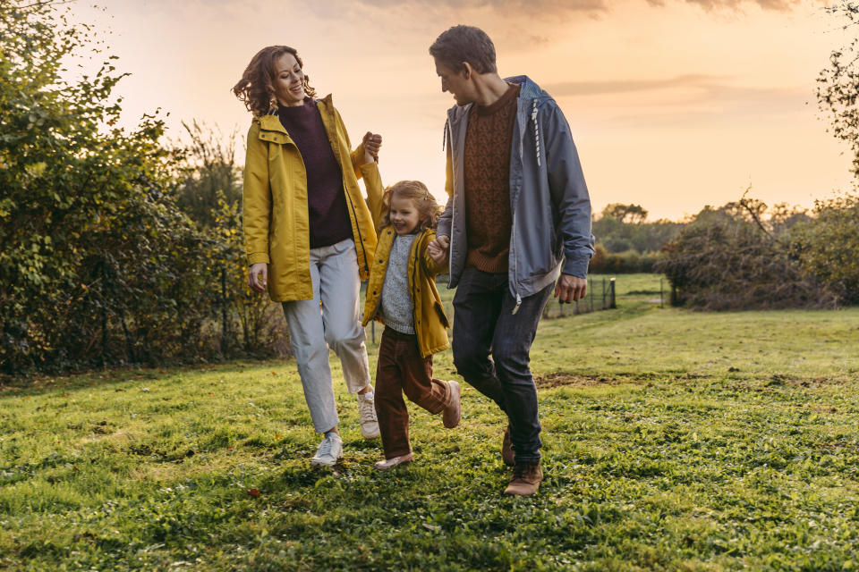 A mother, wearing a yellow raincoat, and a father, wearing a blue hoodie, hold hands with their young daughter between them as they walk through a field in the evening