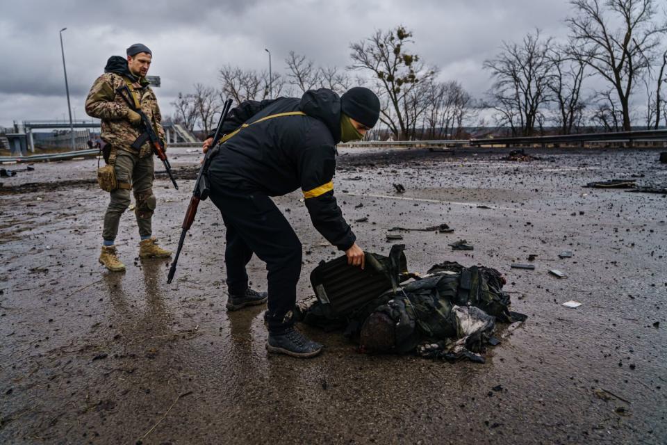 A Ukrainian soldier leans over the body of a Russian soldier while a second Ukrainian stands nearby.