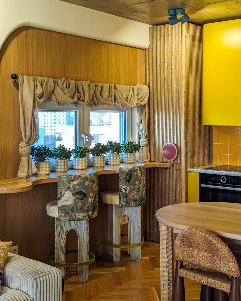 yellow kitchen with wavy wood window counter with botanical fabric backed stools and lots of potted plants