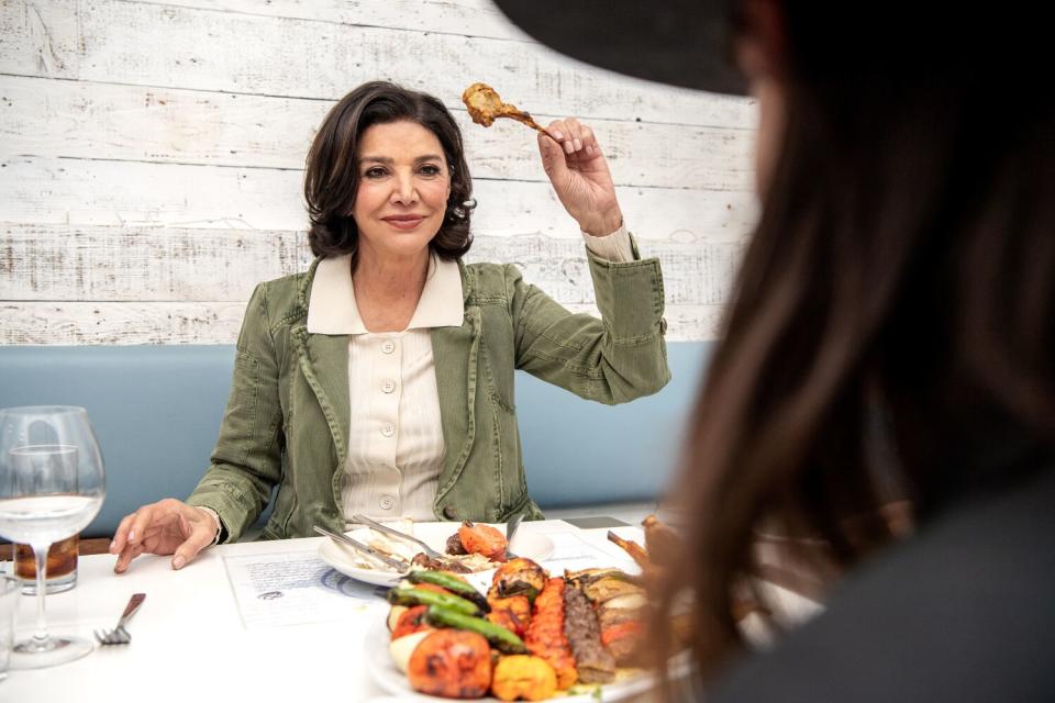 A woman seated at a restaurant table holds up a lamb chop. She's facing another woman with her back to the camera.