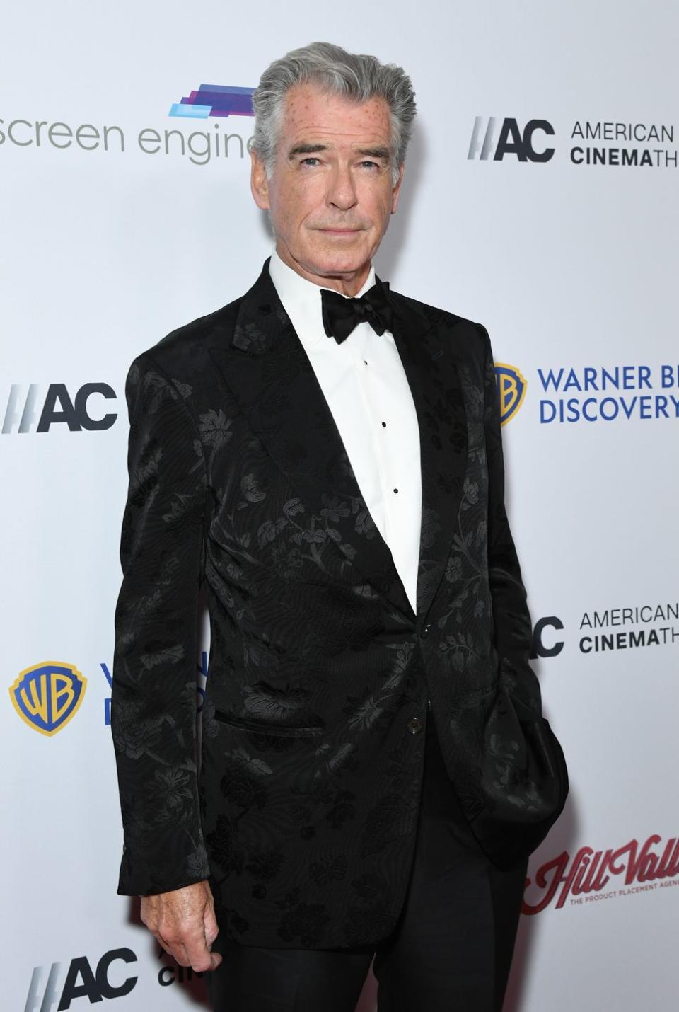 pierce brosnan, an older man stands looking at the camera with a neutral facial expression, he has grey hair and wears a black suit with bow tie