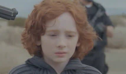 A redhead child looks scared as they stand in a desert with a gun pointed at their head