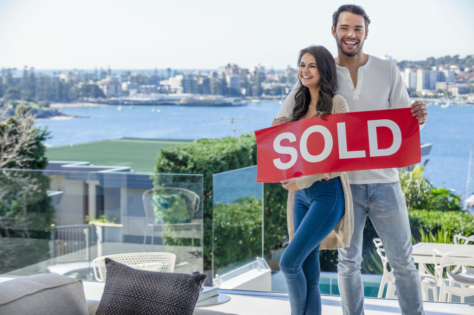 Young couple holding a sold sign in a luxury home. They are standing in the living room, with a view of Sydney harbour in the background. They are both happy and smiling.