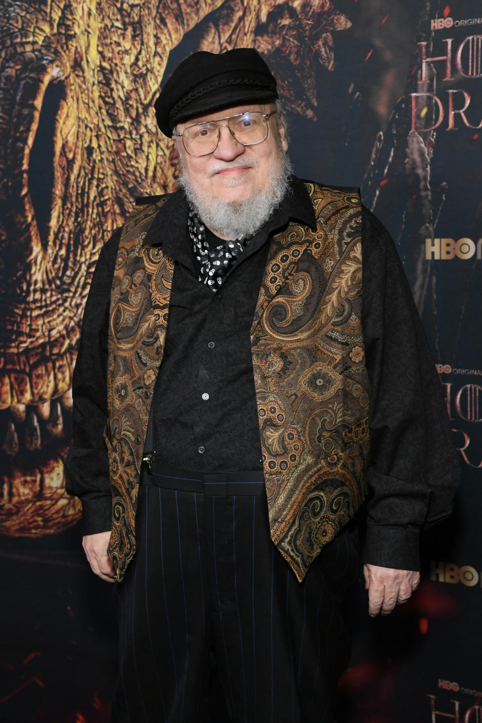 George R.R. Martin on the "House of the Dragon" red carpet, wearing a paisley vest, a dark shirt, striped pants, and a cap