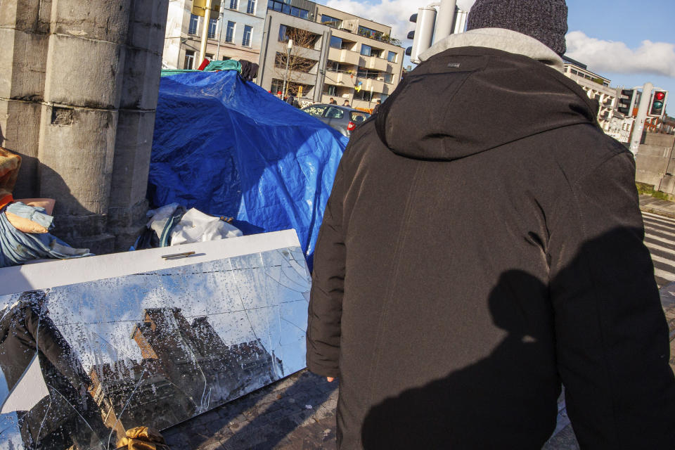 A man walks by a makeshift tent camp as the Petit Chateau reception center is reflected in a mirror in Brussels, Tuesday, Jan. 17, 2023. Many refugees and asylum-seekers are literally left out in the cold for months as the European Union fails to get its migration system working properly. And most talk is about building fences and repatriation instead of working to improve a warm embrace for people fleeing nations like Afghanistan where the Taliban has taken over. (AP Photo/Olivier Matthys)