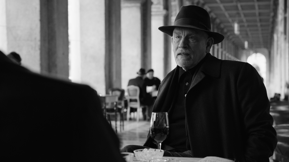 A man in a black 1960s suit and bowler hat sitting at an outdoor table with a glass of wine; still from 'Ripley'