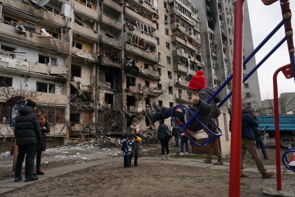 A child on a swing in front of a bombed-out building as residents mill around.