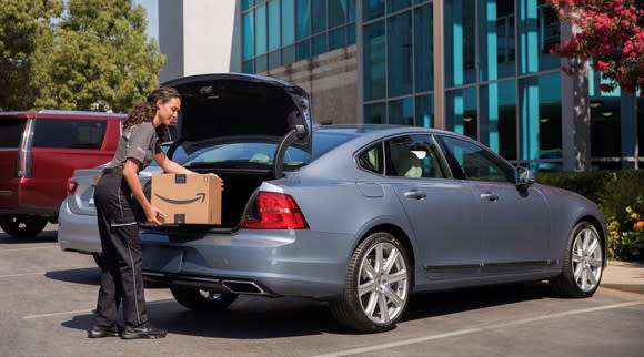 An Amazon delivery person is shown placing a package in the trunk of a 2018 Volvo S90 sedan. The sedan is parked in a corporate parking lot, and its trunk lid is open.