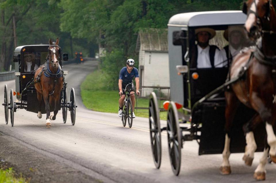 Fernando Maluf rides alongside two horse drawn buggies on Jacksonville Road during the Ironman 70.3 Pennsylvania Happy Valley on Sunday, June 30, 2024.