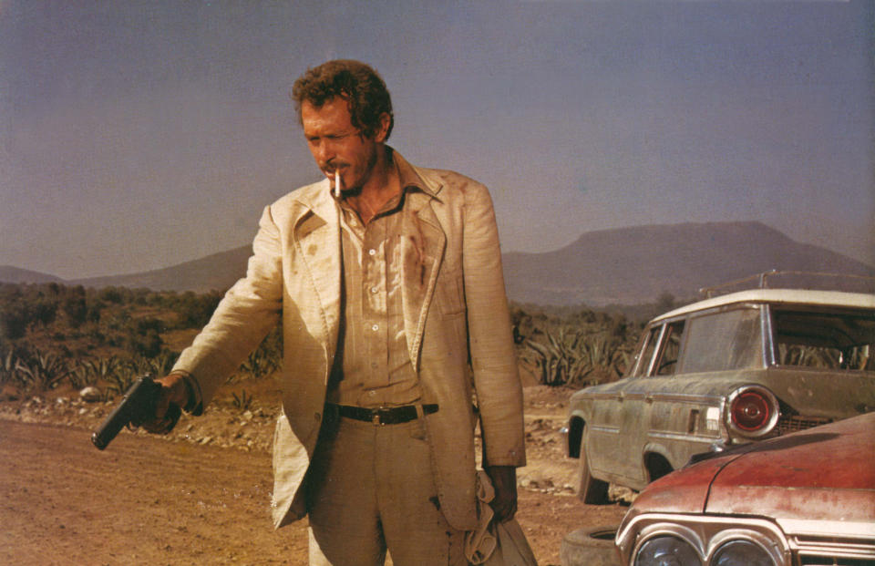 A gun-toting man with a loose cigarette hanging from his mouth stands on a dusty road