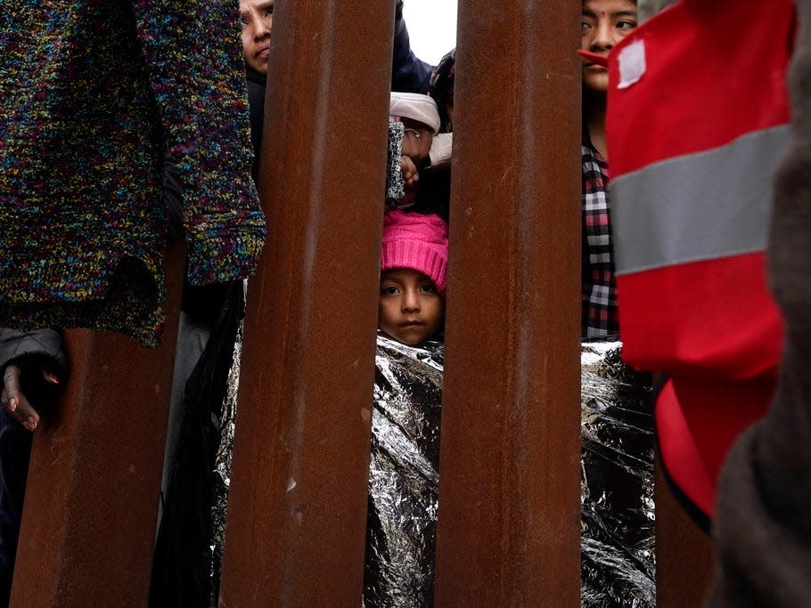 A girl looks on as others reach through a border wall