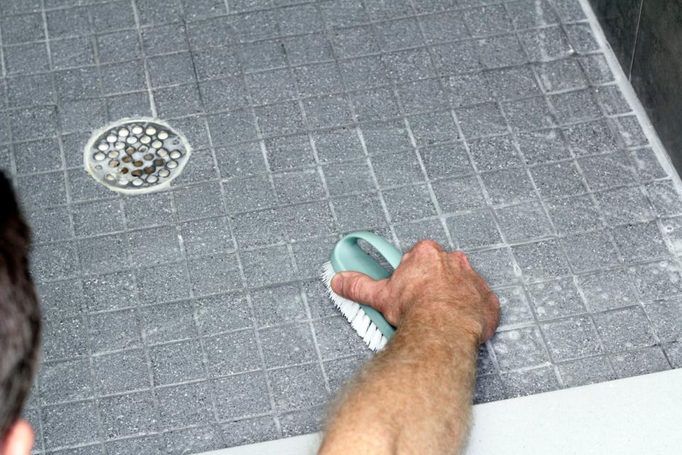 Gray tile floor with grout being cleaned by an adult male with a scrub brush and soap.
