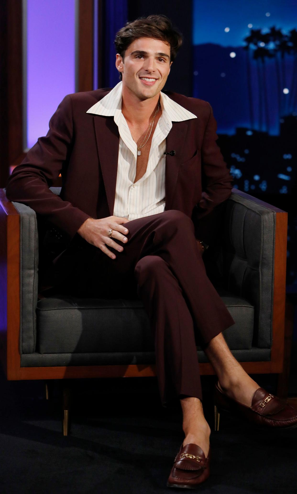 Jacob sitting in a burgundy suit with a white wide-collared and unbuttoned shirt with loafer shoes without socks.