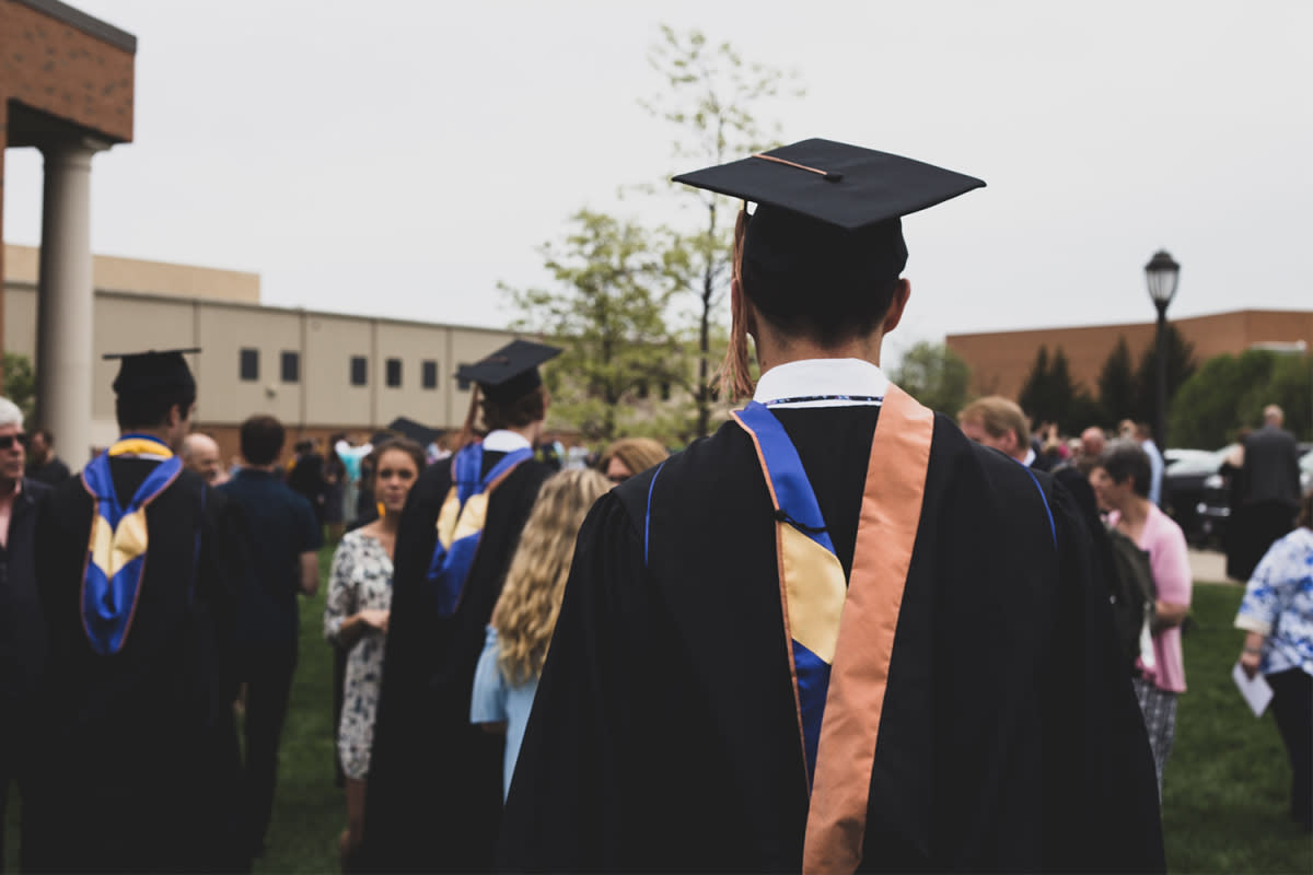 A man facing away from the camera wearing a graduation cap, black robe and stole in the courtyard of a university among other college graduates and family members