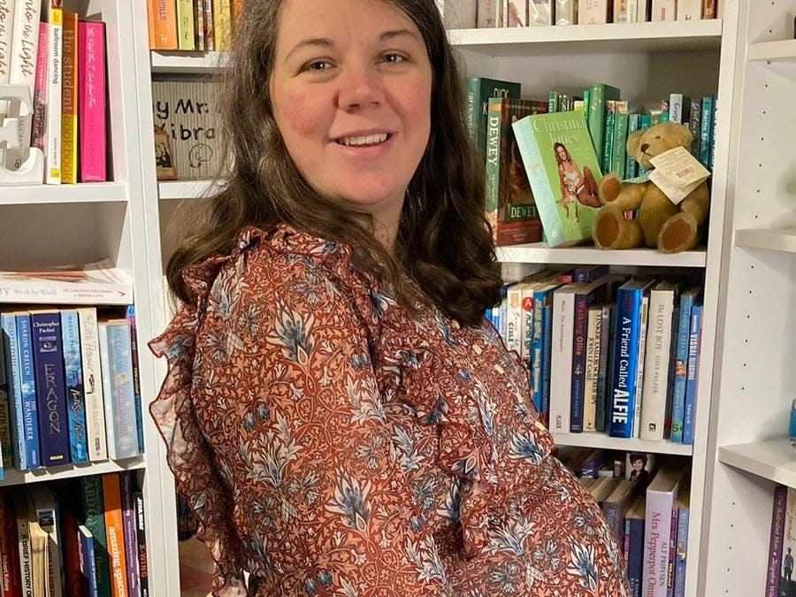 A heavily pregnant woman standing next to a set of bookshelves.