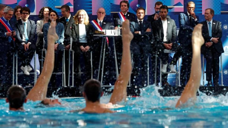 Präsident Macron und andere Beamte beobachten Synchronschwimmer in einem Schwimmbad