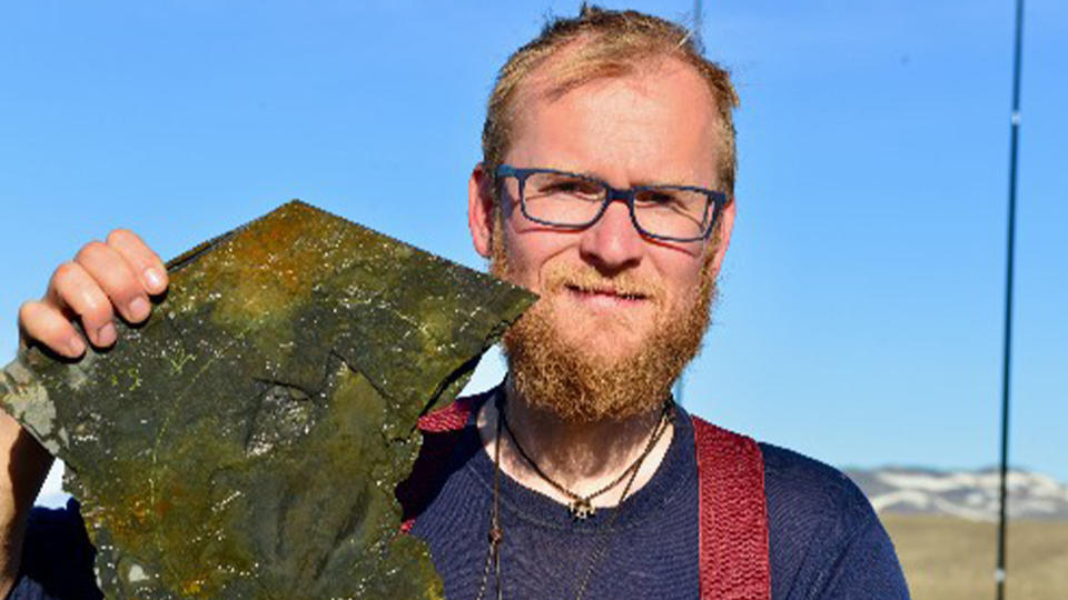 A bearded man holds a fossil against the blue sky.