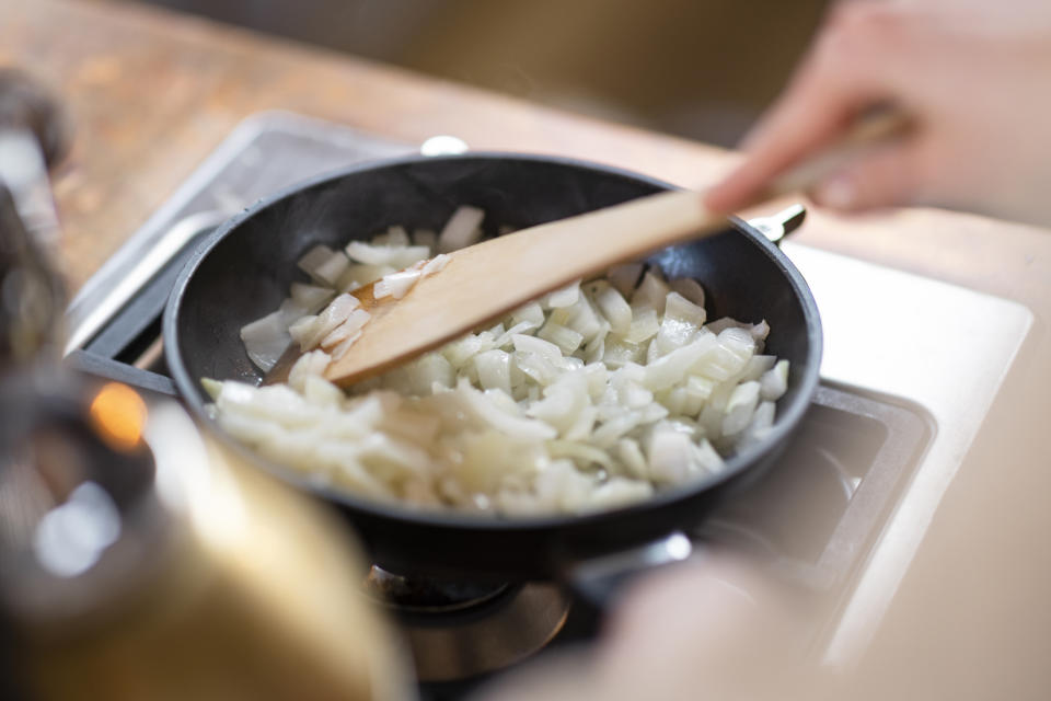 Onions being sautéed in a frying pan, stirred by a wooden spatula held by a person's hand