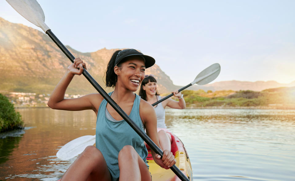 Two women smile while kayaking on a calm river, enjoying the outdoors. They wear casual tank tops and caps. Mountains and greenery are visible in the background