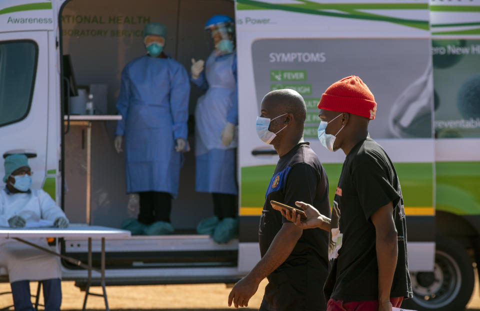 Men walk past health workers in a mobile testing clinic to queue for screening and test for COVID-19 at Lenasia South, south Johannesburg, South Africa, Tuesday, April 21, 2020, during a campaign aimed to combat the spread of Coronavirus. (AP Photo/Themba Hadebe)