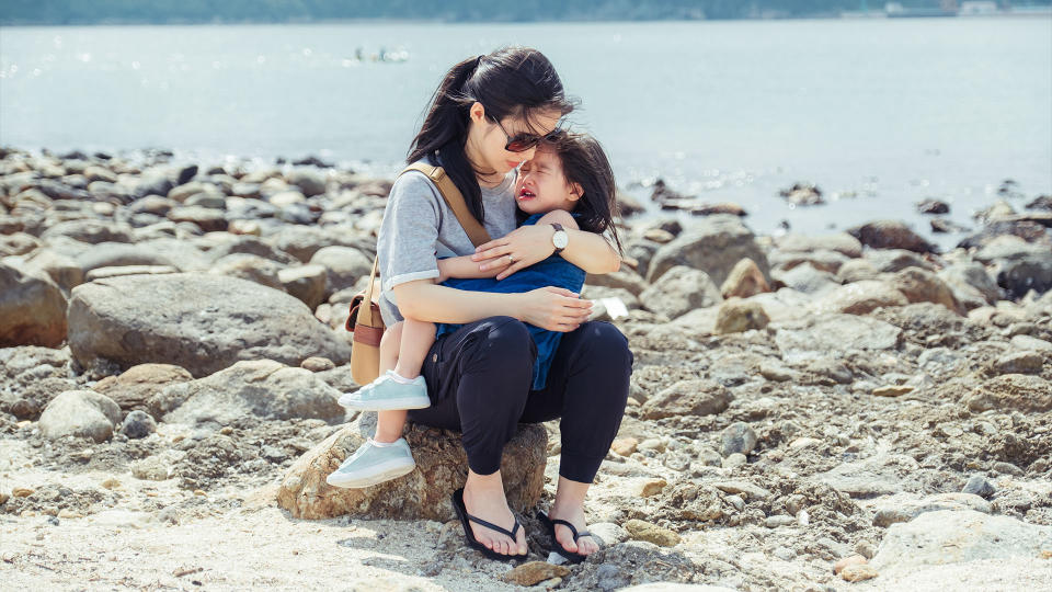 Young mother carrying her crying little daughter in arm who got hurt on the beach on a summer sunny day.