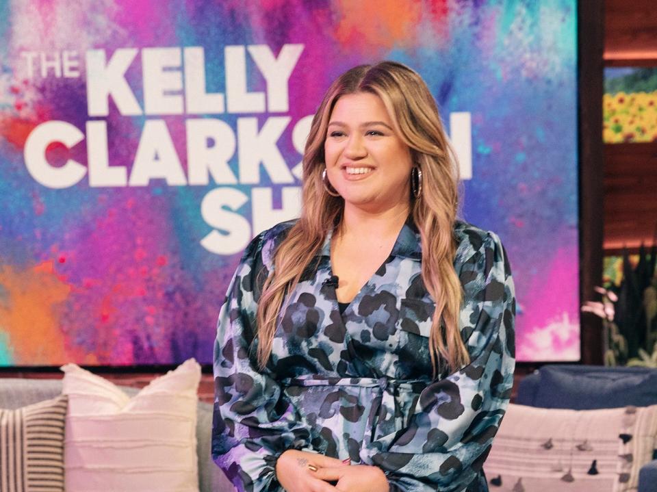 Kelly smiling in a blue dress in front of a Kelly Clarkson backdrop and set.