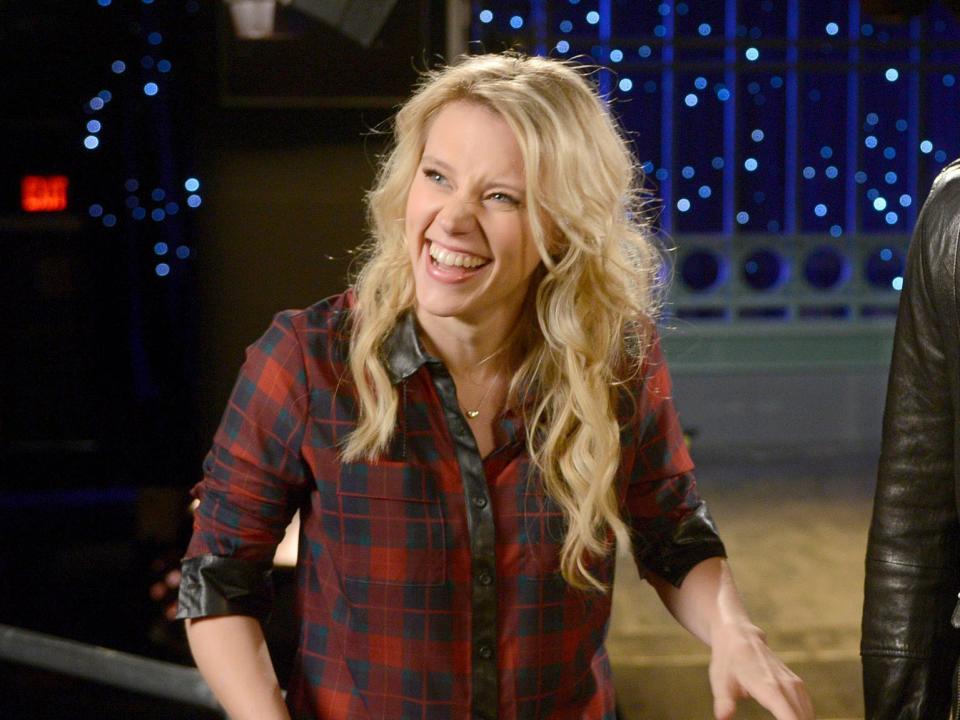 Kate McKinnon with long blonde hair laughing in front of a tv set.