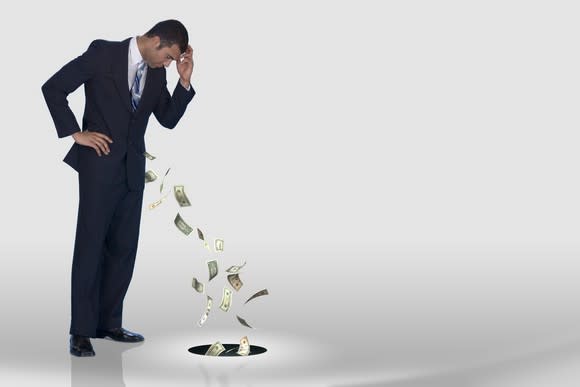 Man in a suit watching money fly out of his pocket and into a hole in the floor