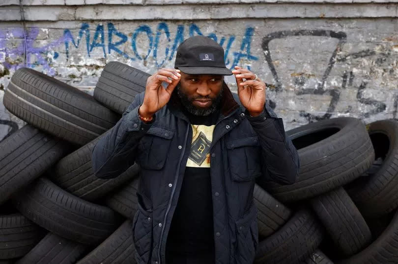 Dwaine Patterson in a jacket and holding his baseball cap in his hands in front of a pile of tyres