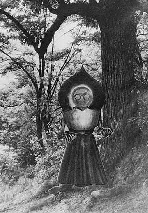 A person stands in a forest wearing a metal suit with a dome-like headgear and large circular eye openings. Trees and foliage are visible in the background