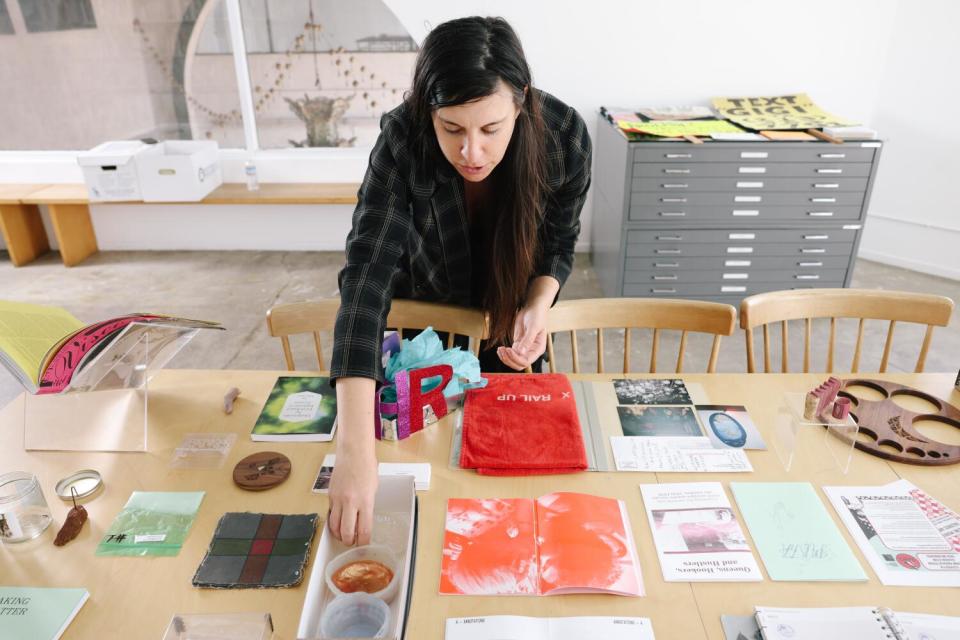 A woman leans over a table covered with artists' materials and works.