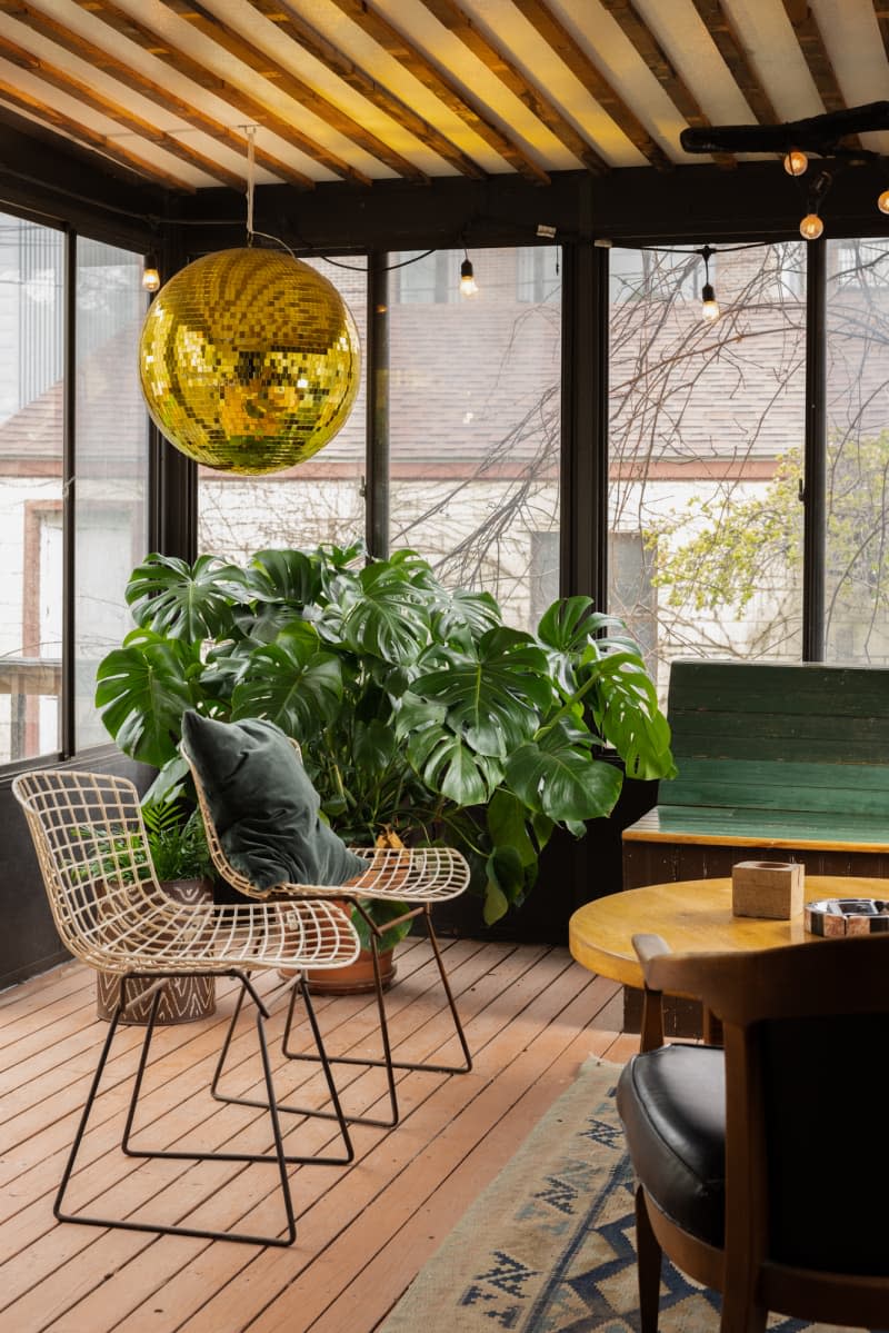 Round gold pendant lamp hanging above wire chairs in sunroom with monstera plants in corner.