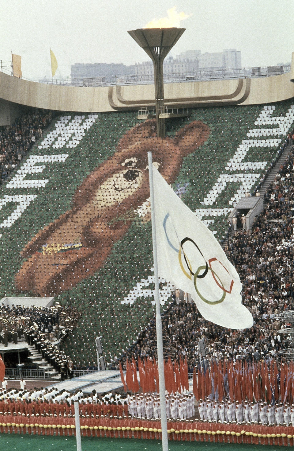 FILE - In this July 19, 1980, file photo, members of the crowd hold up 3,500 cards to create an image of Misha the Bear Cup, the mascot of the Moscow Olympic Games at the Lenin Stadium in Russia on. Above burns the Olympic flame. (AP Photo/File)