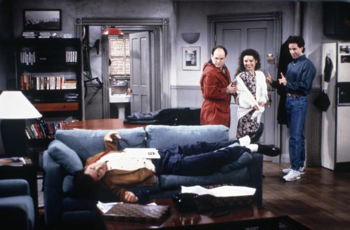Three people stand in the doorway to an apartment and look at a man lying on a couch