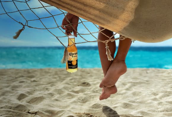 Person holding a bottle of Corona with feet hanging off the side of a hammock.