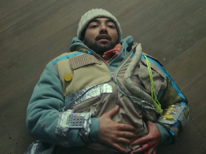 frank wearing a heavy jacket and hat in episode 7 of station eleven, lying on the ground and clutching a bleeding stab wound on his lower left side