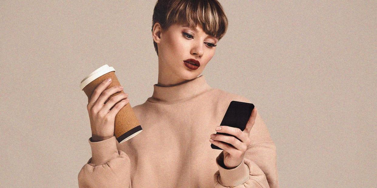 a young woman in a beige jumper in front of a beige background holding a coffee in one hand and a phone in the other