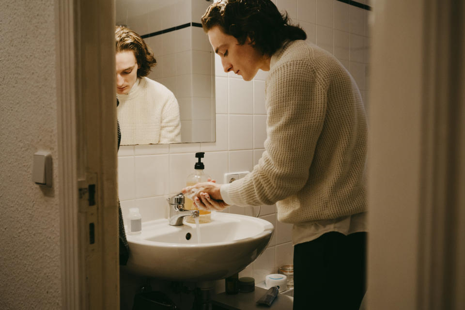 Person in a bathroom washing their hands under a running faucet, with a reflection showing them focused on the task