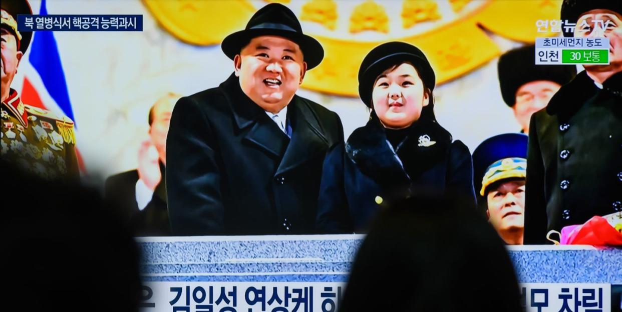 several people watch a television screen, upon which kim jong un and his daughter kim ju ae stand at a podium and look upwards, both wearing black coats and hats