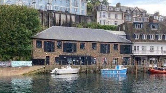 The building in Looe, in Cornwall, which has a banking hub inside is seen across the harbour with boats in front of it, and other buildings high up behind it.