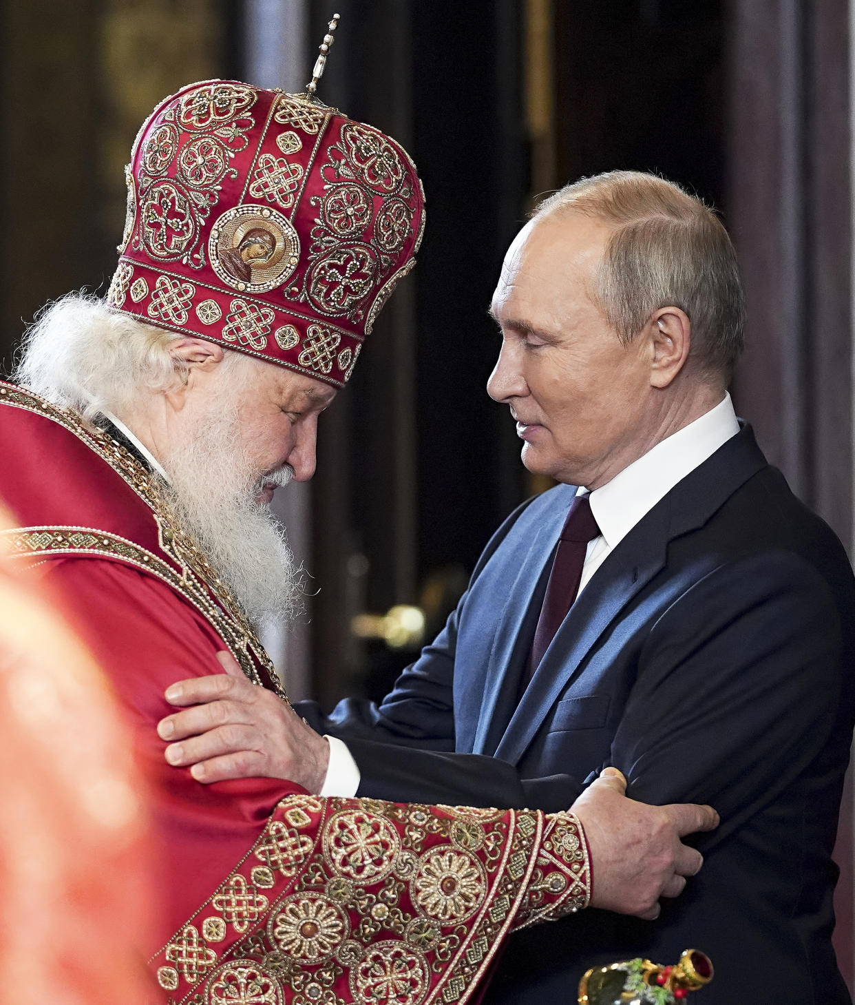 Russian President Vladimir Putin reaches out to put both hands warmly on Patriarch Kirill, who is wearing red robes and miter, and bows his head in a gesture of acknowledgment.