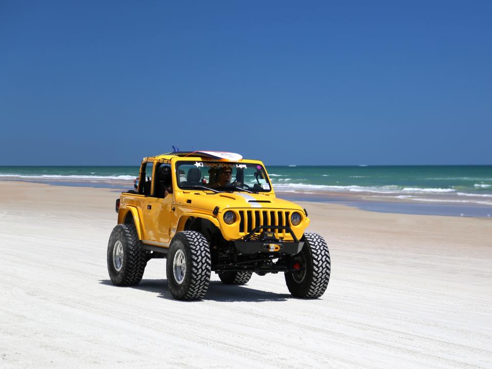 A yellow jeep wrangler with a surfboard on the roof rides on the sand at Daytona Beach.
