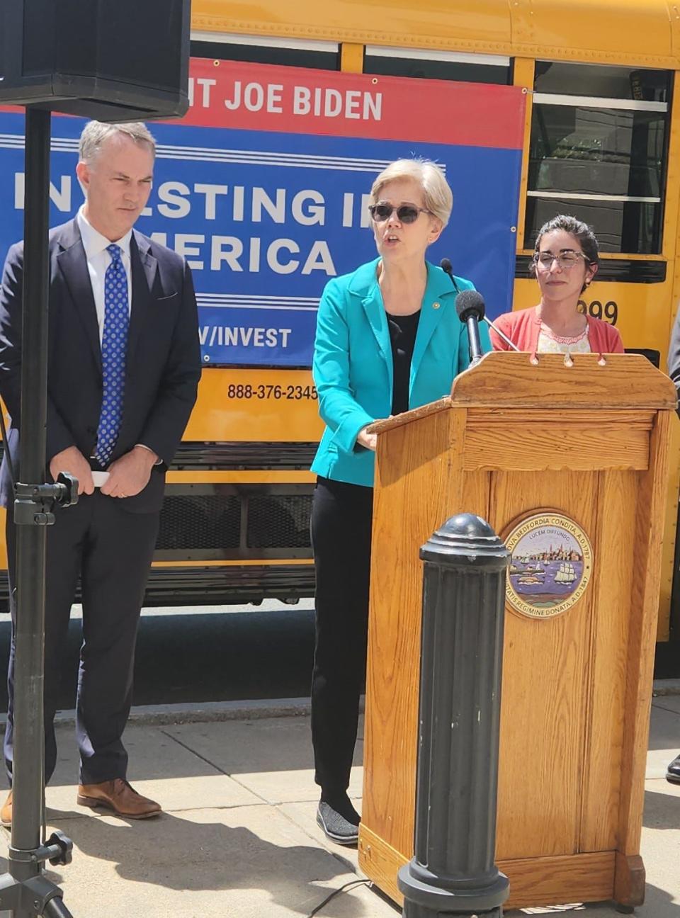 U.S. Senator Elizabeth Warren speaks to the crowd outside of Roosevelt Middle School on Friday, May 26, as one of New Bedford's new electric school buses can be seen in the background. Warren is seen here flanked by Mayor Jon Mitchell (left) and Dr. Amelia Monteiro (right).