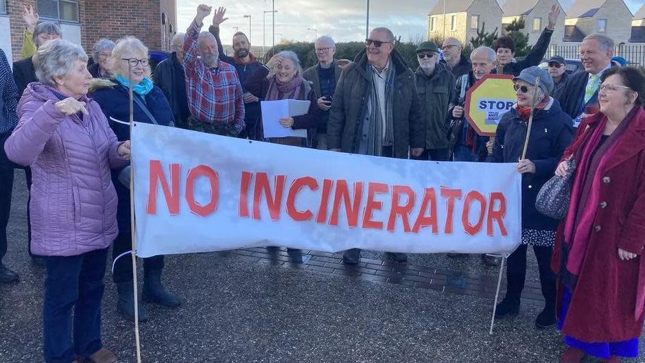 A group of about 15 people are standing with a banner that says: "no incinerator" in red, capital letters on a white background