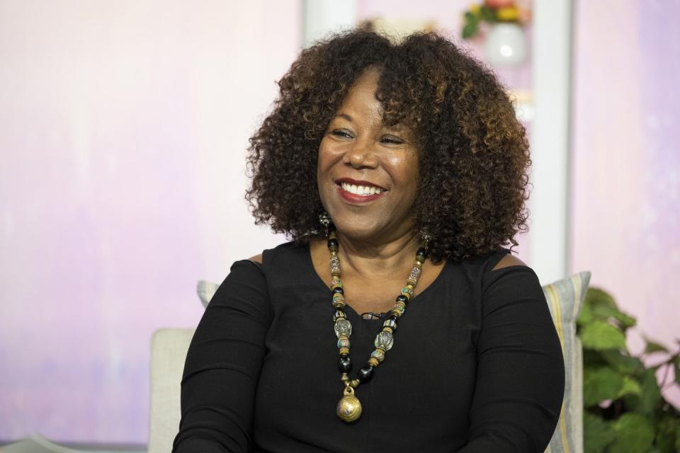 ruby bridges smiles and looks left, she wears a dark long sleeve shirt with a large beaded necklace, she sits in a chair with a pillow behind her