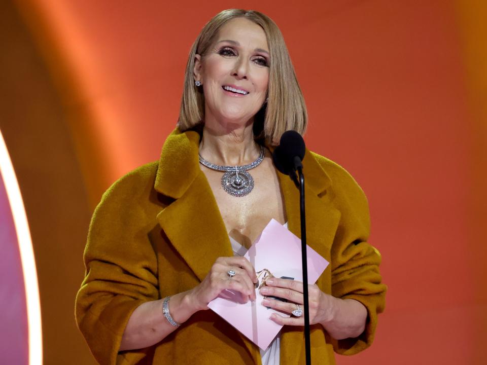 celine onstage at the grammy awards, standing in front of a microphone holding a pink envelope and wearing a brown overcoat