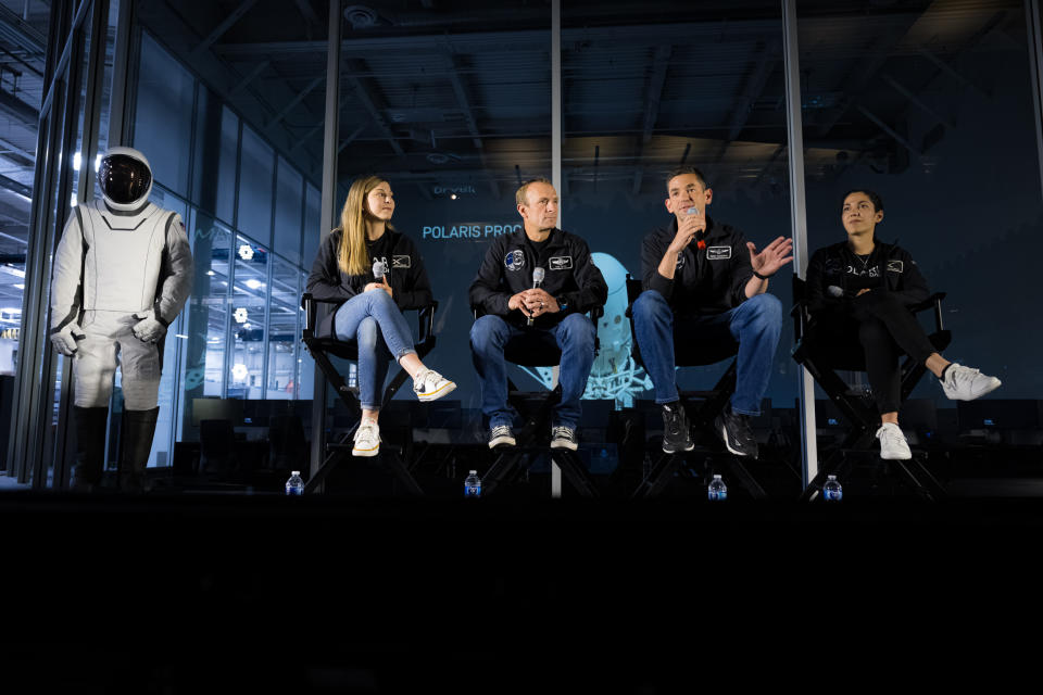 Four people in black coats sit in high chairs in front of a wall of glass.  To their left is a white space suit.