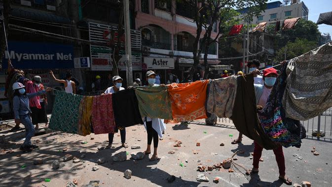 Perempuan menggantung pakaian tradisional Myanmar, longyi, di seberang jalan selama demonstrasi menentang kudeta militer di Yangon, Senin (8/3/2021). Jemuran kain itu untuk membatasi gerak polisi dan tentara karena berjalan di bawah jemuran pakaian ini dianggap akan membawa sial bagi pria. (STR/AFP)