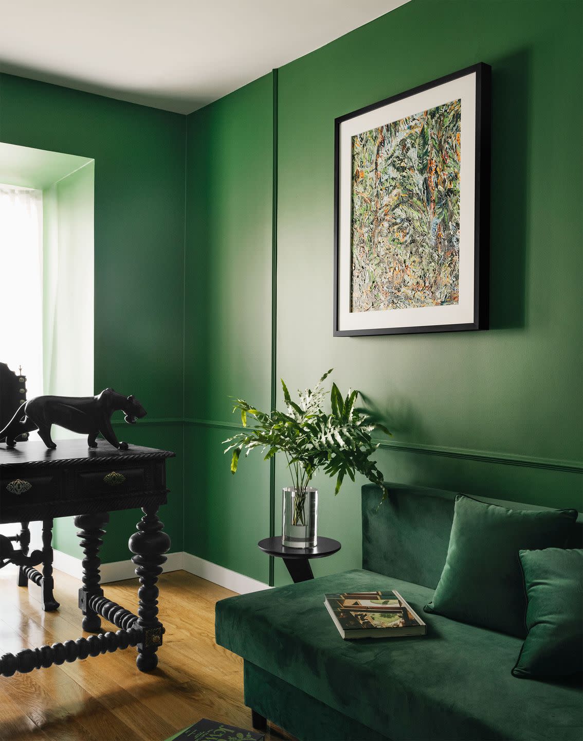 a home office with deep emerald walls has an emerald velvet sofa, a small black side table with a vase of ferns, a dark wood desk with ornate legs and a panther on top, and a framed abstract painting